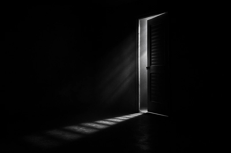 KI-generiertes Bild. Eine halb geöffnete Tür lässt einen schmalen Lichtstrahl in einen dunklen, leeren Raum fallen. Das Licht durchbricht die Schatten und erzeugt eine ruhige, nachdenkliche Stimmung – ein Sinnbild für Orientierung in der Dunkelheit.