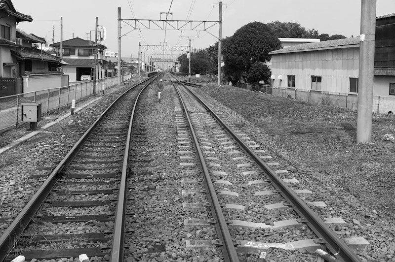 Schwarz‑weiß‑Foto zweier parallel verlaufender Eisenbahnschienen, die sich in der Ferne perspektivisch verjüngen. Links und rechts der Gleise stehen japanische Wohnhäuser, Zäune, Strommasten und niedrige Industriegebäude. Über den Gleisen verlaufen Oberleitungen, und am Horizont verschwimmen die Strukturen in der Ferne.