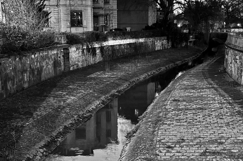 Schwarz‑weiß‑Fotografie eines schmalen Wasserkanals, der zwischen zwei gepflasterten Wegen verläuft. Die linken und rechten Ufer werden von alten Steinmauern eingefasst, teilweise mit Graffiti bedeckt. Im Hintergrund stehen mehrstöckige Gebäude, Bäume und ein kleiner Brückenbogen, während sich einzelne Fenster und Strukturen im ruhigen Wasser spiegeln.